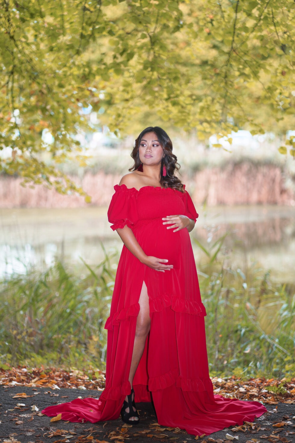 Een zwangere vrouw in een lange rode jurk staat in een veld, met groen en roze bloemen op de achtergrond.