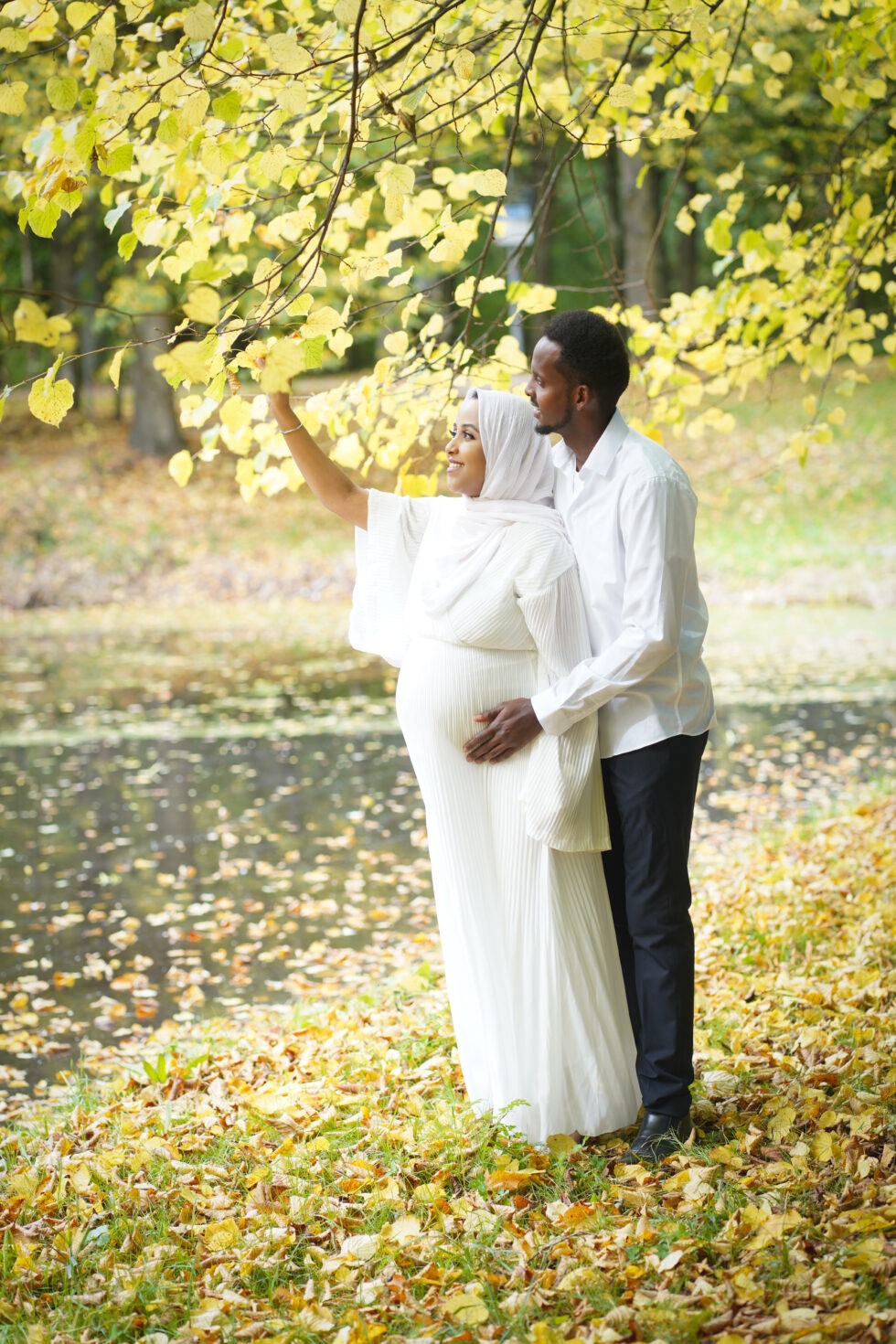 Een stel staat bij een vijver in een herfstpark, met de vrouw in een witte jurk.