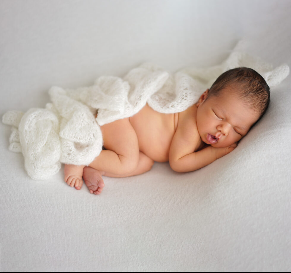 Pasgeboren baby in een witte doek met een zachte bontmuts, liggend op zijn rug op een donzige crèmekleurige deken