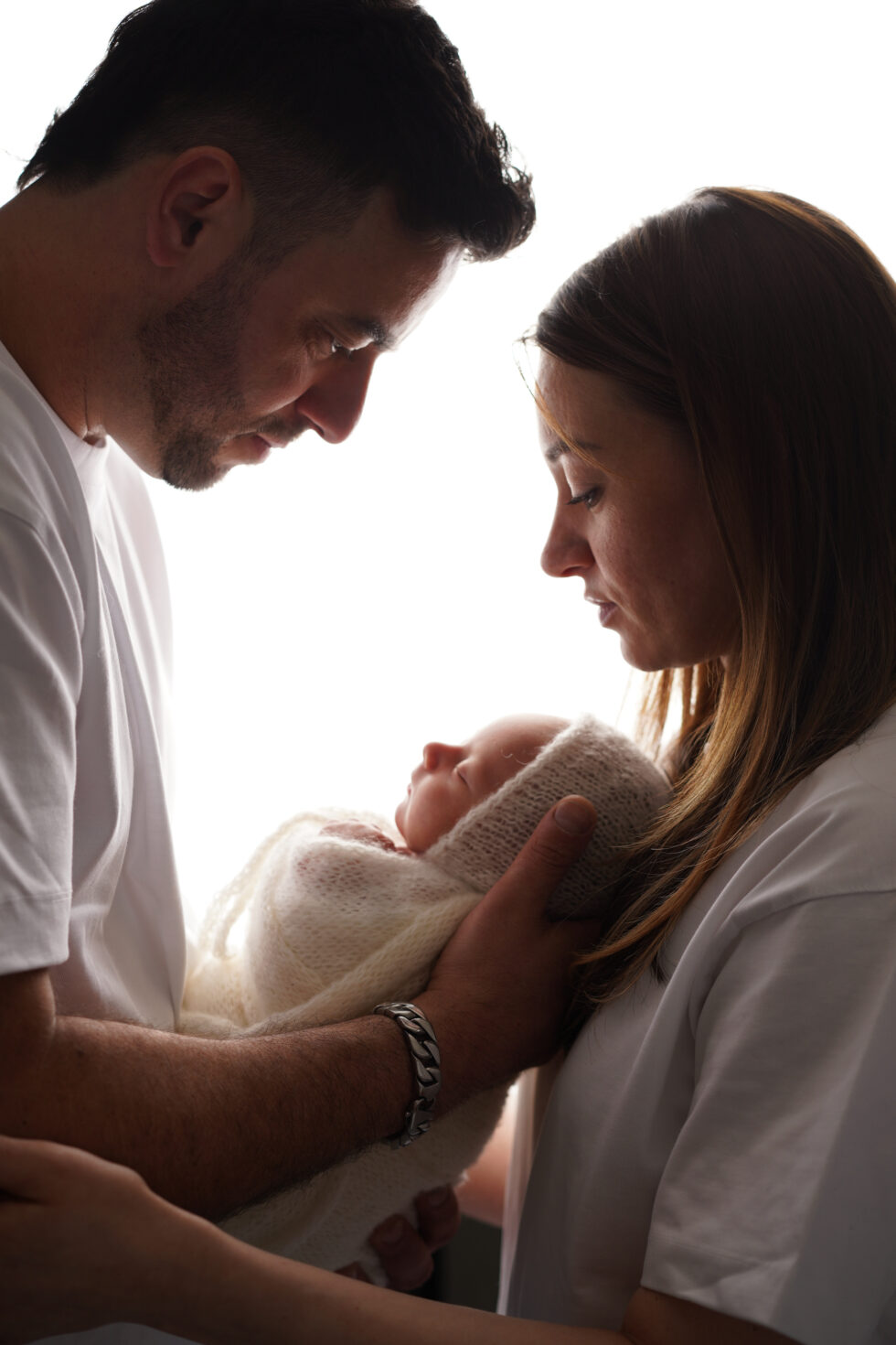 Silhouet van ouders die zachtjes naar hun pasgeboren baby kijken, met zacht licht dat hun gezichten benadrukt.