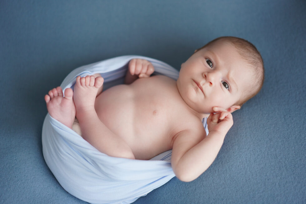 Een baby ligt op een blauwe achtergrond, omwikkeld met een witte doek.