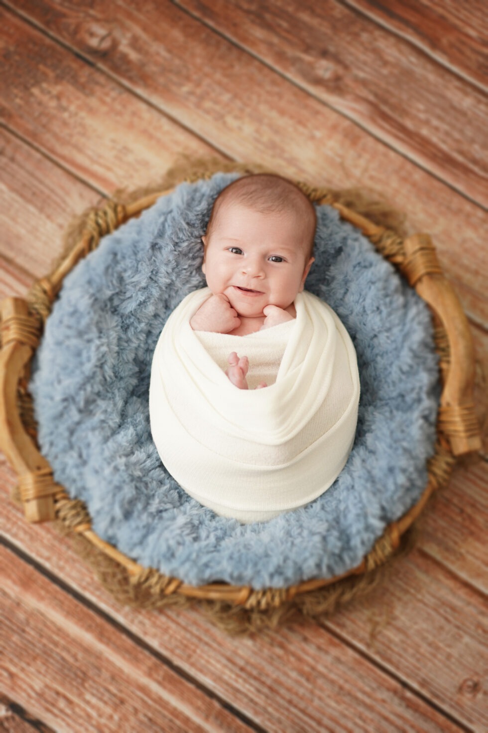 Pasgeboren baby in een witte doek, glimlachend naar de camera terwijl hij op een donzig blauw kussen ligt in een ronde houten mand