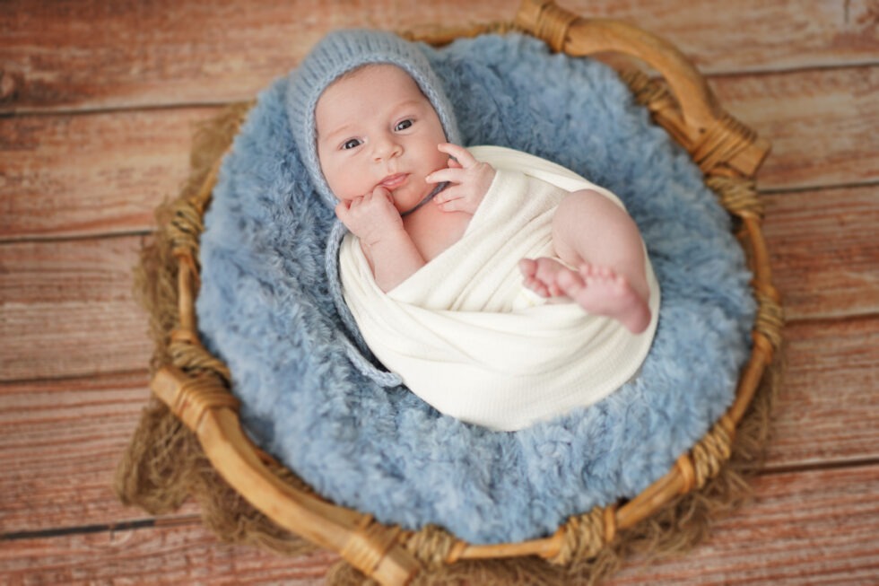 Pasgeboren baby in een witte doek, glimlachend naar de camera terwijl hij op een donzig blauw kussen ligt in een ronde houten mand.