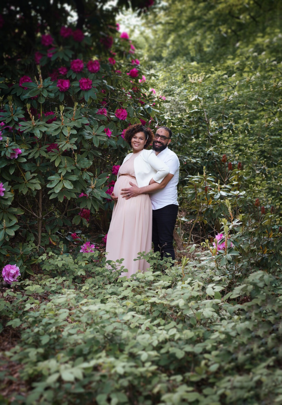 Een stel staat tussen bloeiende bloemen, met de vrouw in een roze jurk.