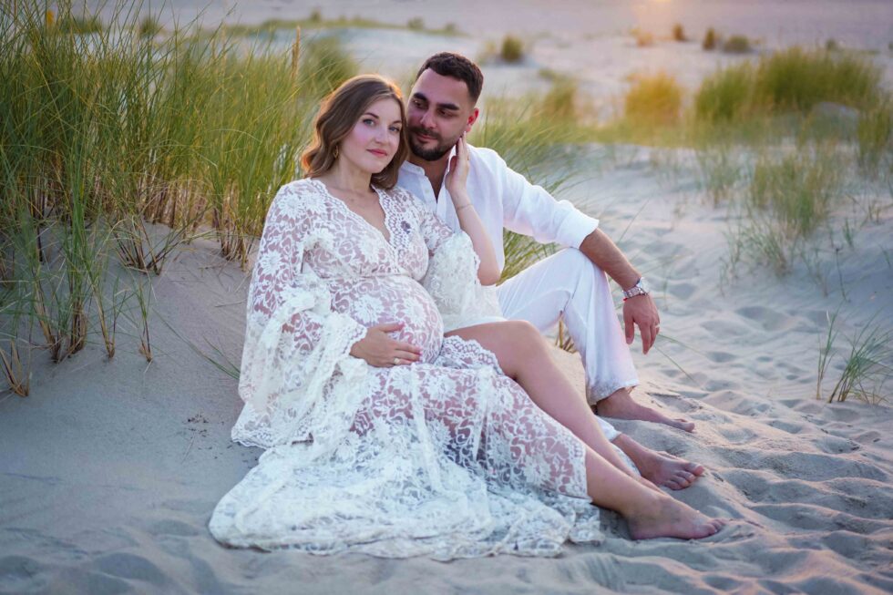 Een stel zit op het zand bij het strand, met de vrouw in een zwierige witte jurk.