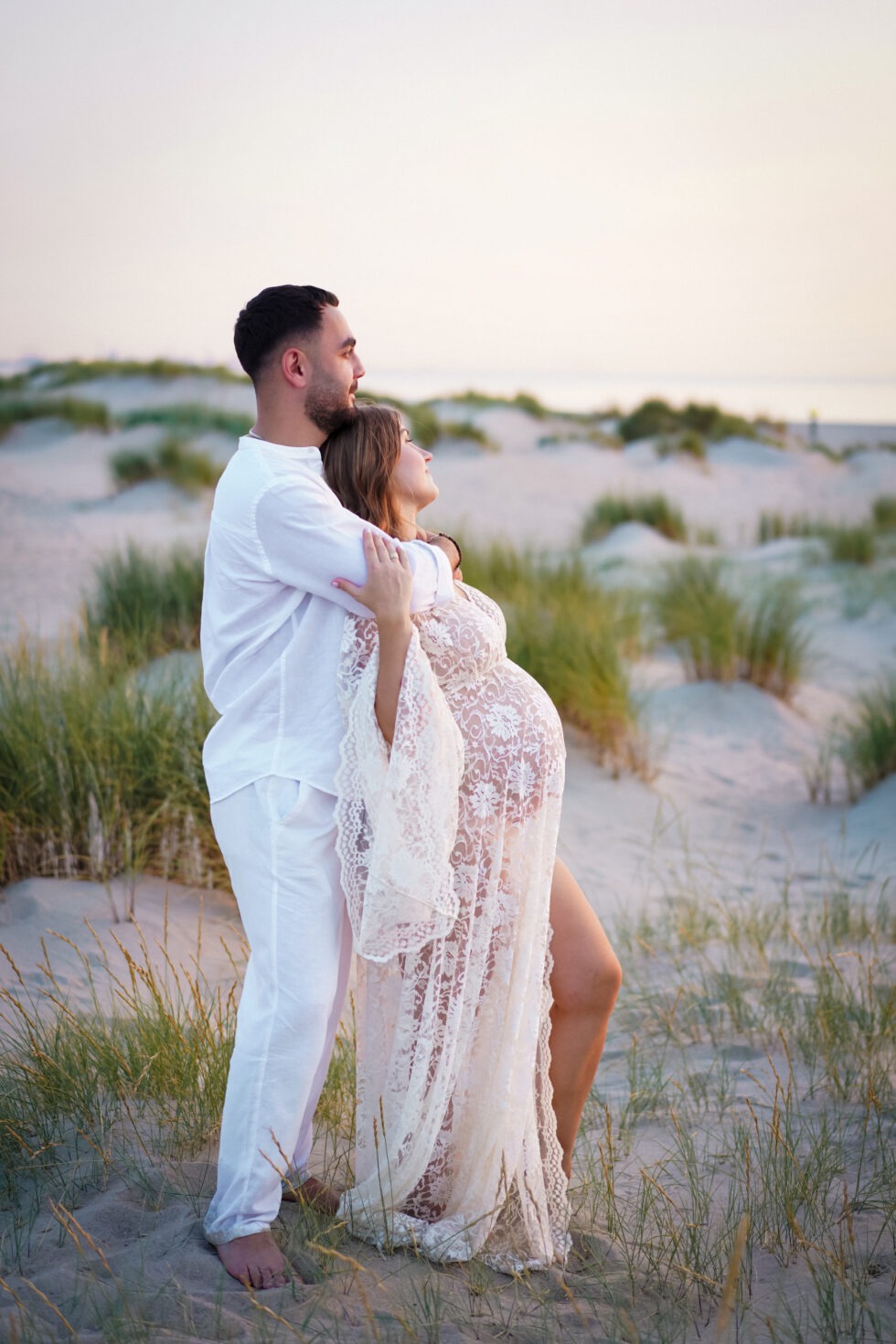 Een zwangere vrouw in een witte kanten jurk staat in een grasrijk duingebied, terwijl ze haar buik vasthoudt en haar partner haar van achteren omhelst