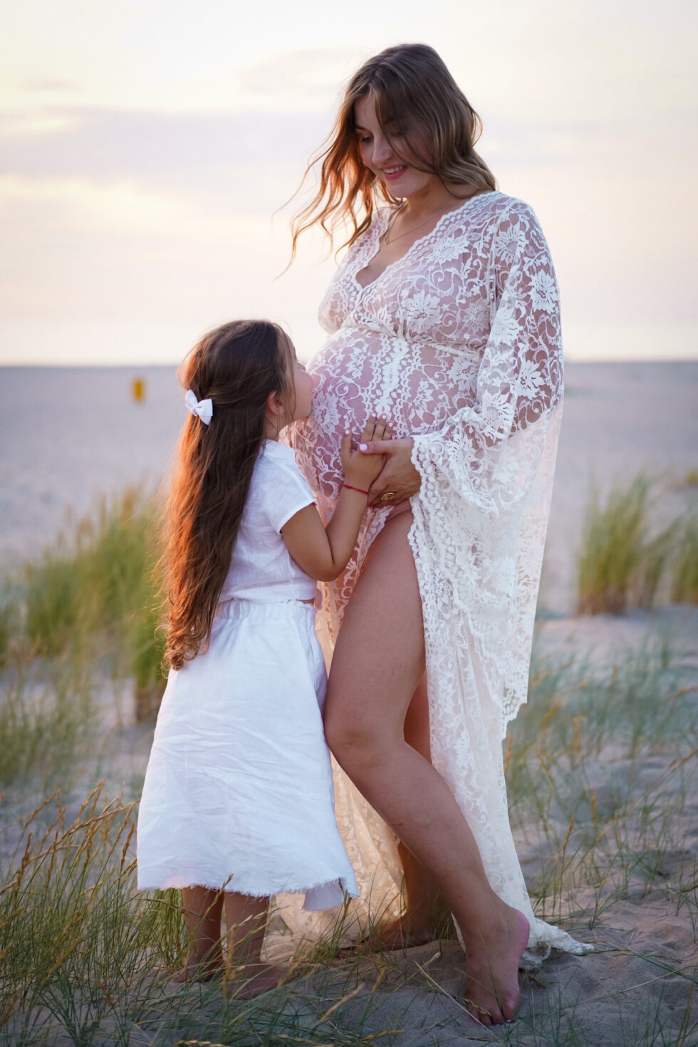 Een jonge dochter kust de zwangere buik van een vrouw in een zwierige witte jurk, terwijl ze samen op het zand bij het strand zitten.