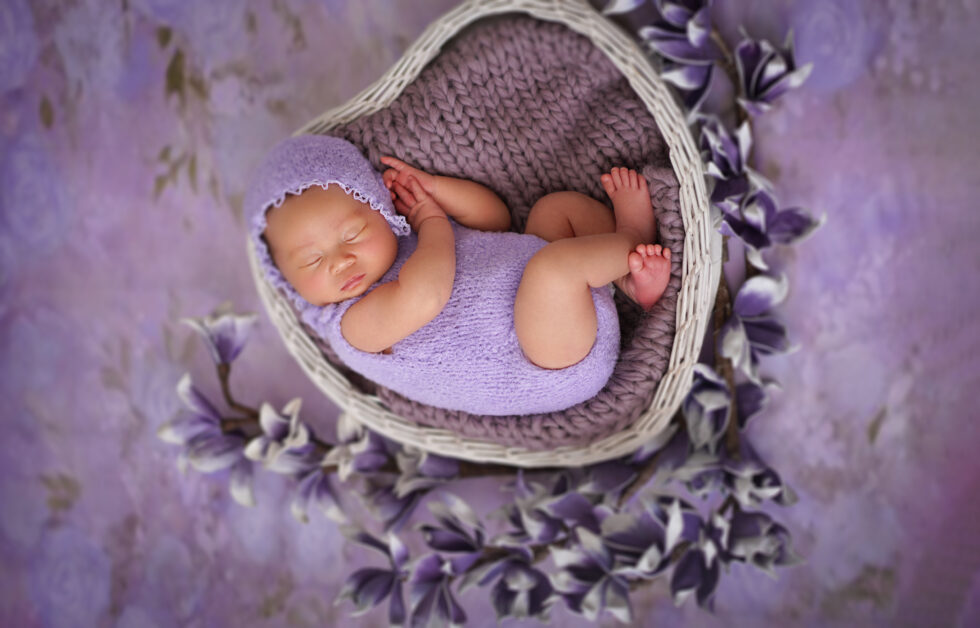 Pasgeboren baby rustend in een hartvormige mand, omringd door een paarse en grijze deken.
