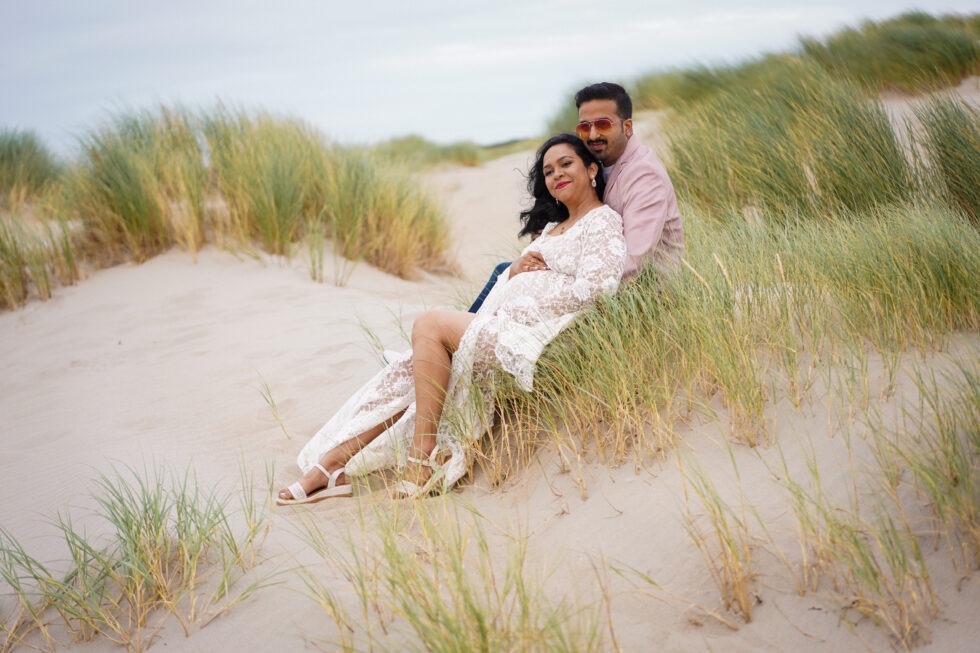 Een zwangere vrouw in een witte kanten jurk zit in een grasrijk duingebied, terwijl ze haar buik vasthoudt en haar partner haar van achteren omhelst.