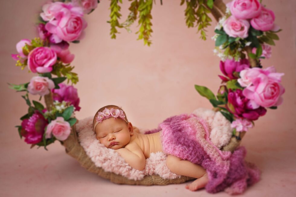 Pasgeboren meisje in een roze bloemenjurk, liggend in een rustiek houten bed, omringd door roze en witte bloemen.