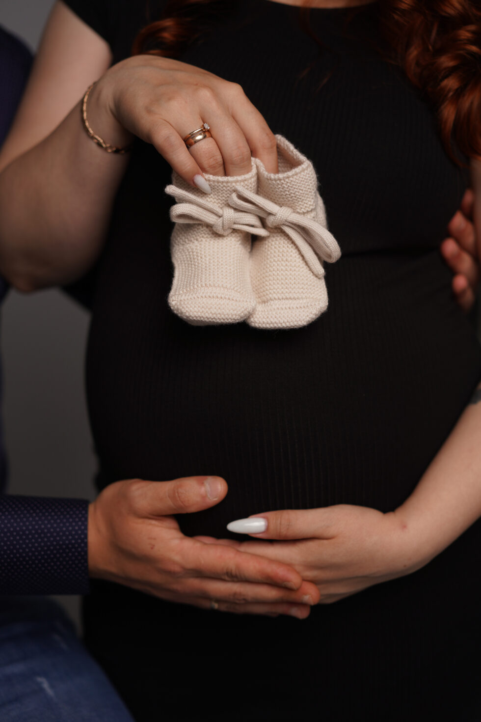 Een close-up van een zwangere buik in een zwarte jurk, met de handen van het stel die voorzichtig een paar babyschoentjes vasthouden.