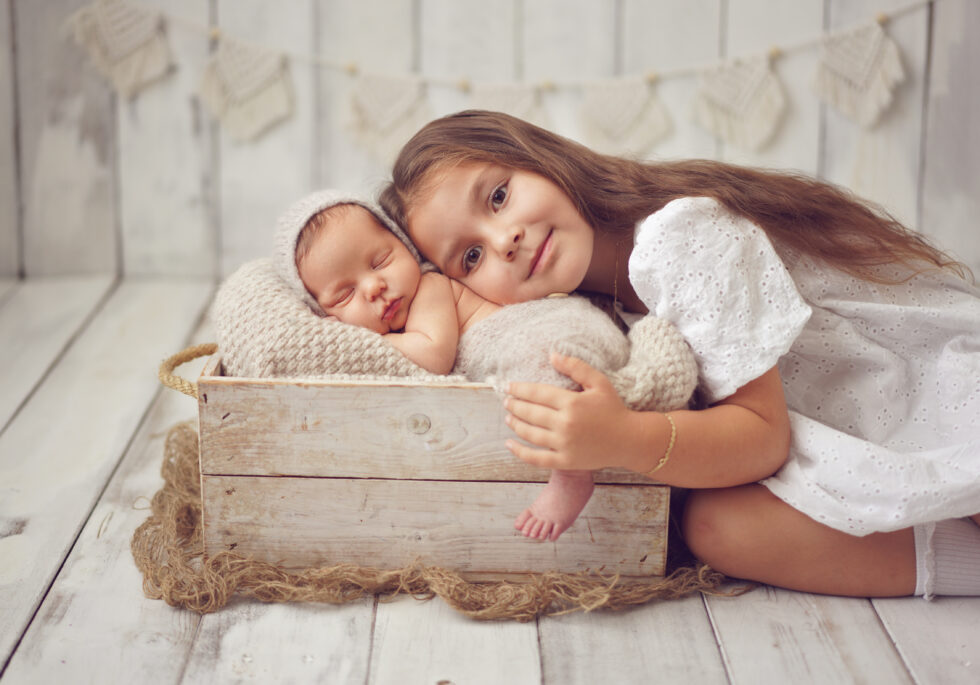 Pasgeboren baby slapend in een houten doos, met een oudere zus die haar zachtjes knuffelt.
