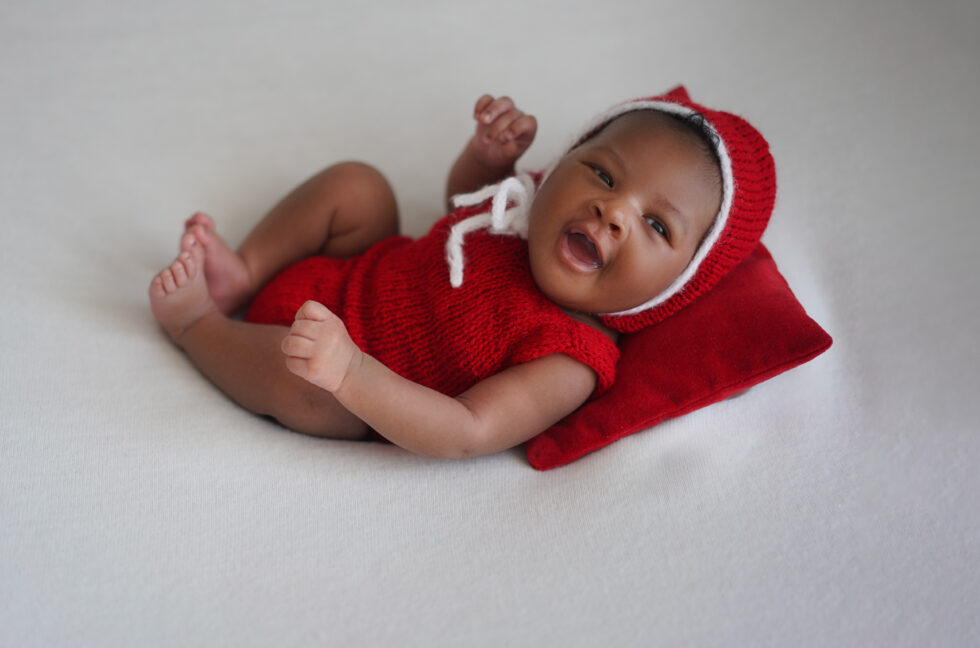 Pasgeboren baby in een rood rompertje, die omhoog kijkt terwijl hij op zijn rug ligt op een witte deken