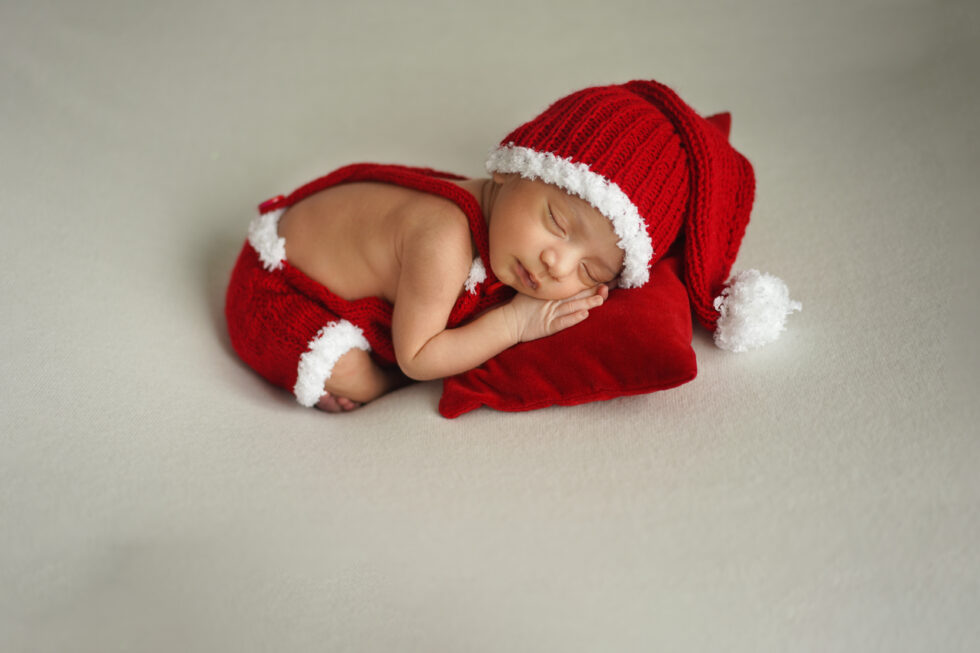 Pasgeboren baby in een rood outfit met een bijpassende hoed, liggend op zijn rug op een zacht rood kussen.